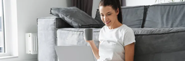 笑顔の女性はコーヒーを飲みながら 自宅で床に座っている間にラップトップを使用して — ストック写真