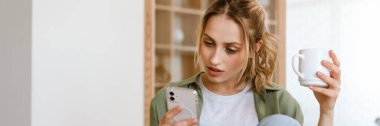 Genç beyaz kadın evde kahve içerken akıllı telefon kullanıyor.