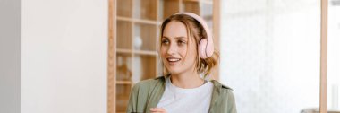 Evde cep telefonu ve kulaklıkla müzik dinlerken gülümseyen genç bir kadın.