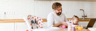 Beyaz baba evde dizüstü bilgisayar kullanırken kızıyla gülüp oynuyor.
