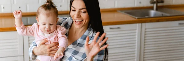 若い母親は自宅でキッチンでラップトップで働いている間ジェスチャーし 彼女の赤ちゃんを保持 — ストック写真