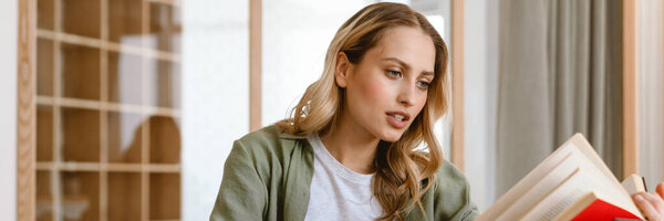 Pensive young white blond woman writing down notes while sitting at wooden table at home holding book