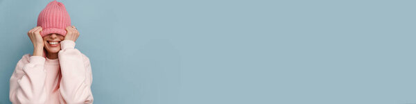 Молодая белая женщина с розовыми волосами улыбается и тянет шляпу вниз на ее лицо изолированы над синей стеной