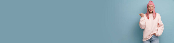 Молодая женщина в розовой шляпе смеется и указывая пальцем в сторону изолированы над синей стеной