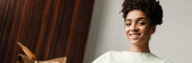 Young black woman smiling and drinking coffee while sitting on armchair at home