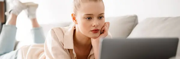 年轻的金发女人坐在现代客厅里 躺在沙发上 一边用笔记本电脑 — 图库照片