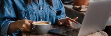 Kafe masasında kahve içerken dizüstü bilgisayar kullanan gülümseyen Afrikalı genç bir kadın, çalışıyor ya da çalışıyor.
