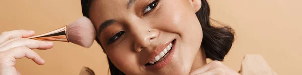 若いですアジアのブルネット女性笑顔ながら使用してパウダーブラシ隔離上ベージュ背景 — ストック写真
