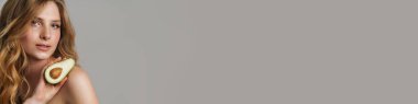 Avokadoyla poz veren yarı çıplak, beyaz bir kadın gri arka planda izole edilmiş.