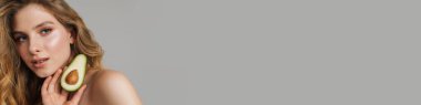 Avokadoyla poz veren yarı çıplak, beyaz bir kadın gri arka planda izole edilmiş.