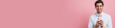 Genç beyaz adam papyon takıyor gülümsüyor ve pembe arka planda cep telefonu kullanıyor.