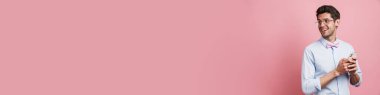Genç beyaz adam papyon takıyor gülümsüyor ve pembe arka planda cep telefonu kullanıyor.