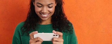 Mutlu gülümseyen Afrikalı sıradan esmer kadın turuncu duvar arkasında duruyor, cep telefonunu tutuyor, oyunlar oynuyor, video izliyor.