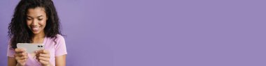 Mor arka planda izole edilmiş, gülen ve cep telefonu kullanan genç siyah bir kadın.