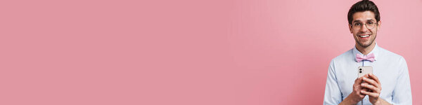 Молодой белый мужчина в галстуке-бабочке улыбается и использует мобильный телефон изолирован на розовом фоне