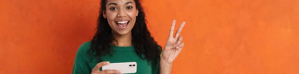 快乐的微笑着的非洲黑发女人 站在橙色墙的后面 拿着手机 玩游戏 看视频 — 图库照片