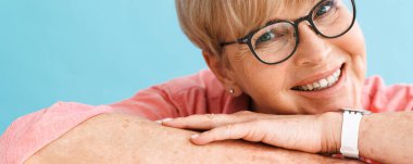 Gözlüklü Avrupalı olgun bir kadın gülümserken mavi arka planda tek başına masaya yaslanıyor.