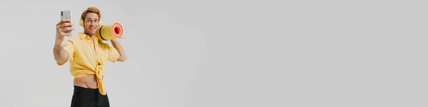 Улыбающийся молодой белый гей с ковриком для фитнеса слушает музыку с наушниками, стоящими на фоне серой стены, делая селфи