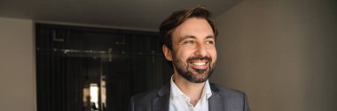 Resmi olarak gülümseyen orta yaşlı beyaz adam otel odasında elinde fincanla dikiliyor.