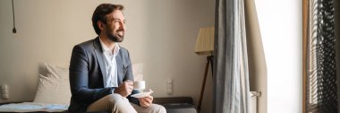 Resmi olarak gülümseyen, orta yaşlı, sakallı bir adam otel odasında bir fincan kahve içerken giyiniyor.
