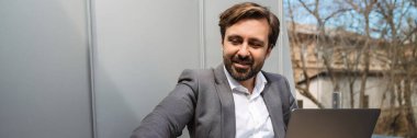 Gülümseyen orta yaşlı sakallı iş adamı balkonda oturuyor dizüstü bilgisayarla cep telefonuna uzanıyor.