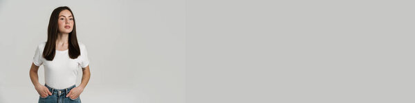 Крупный план портрета молодой белой женщины с длинными каштановыми волосами в белой футболке, стоящей на сером фоне, глядя в сторону