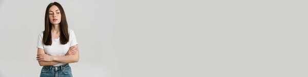 Close up portrait of a young white woman with long brown hair wearing white t-shirt standing over gray background arms folded looking aside