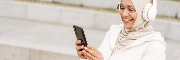 Smilende Middelaldrende Muslimsk Kvinne Hodetelefoner Som Holder Mobiltelefon Stående Gate – stockfoto