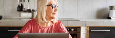 Dizüstü bilgisayarla çalışan, gözlüklü yaşlı bir kadın hafif mutfağın kenarına bakıyor.