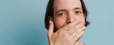 Ağzını sakallı genç kızıl adam mavi arka planda izole edilmiş kameraya bakıyor.