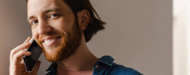 Kapalı alanda cep telefonuyla konuşurken gülümseyen sakallı genç kızıl adam.