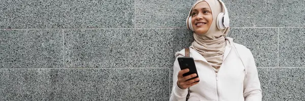 Leende Medelålders Muslimsk Kvinna Hörlurar Med Mobiltelefon Lutad Mot Vägg — Stockfoto