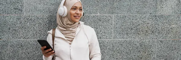 Smilende Middelaldrende Muslimsk Kvinne Hodetelefoner Som Holder Mobiltelefonen Lenende Vegg – stockfoto