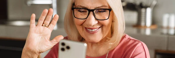ライトキッチンで携帯カメラを振っているメガネの笑顔の高齢女性 — ストック写真