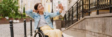 Esmer kadın, şehirde tekerlekli sandalyede otururken cep telefonuyla selfie çekiyor.