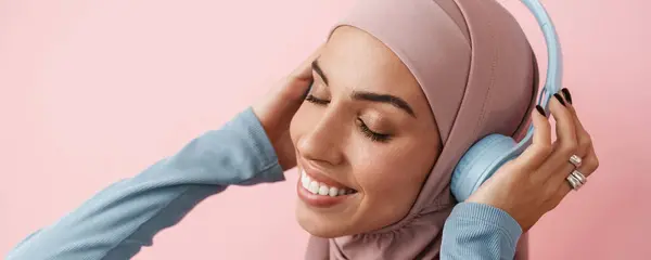 Muotokuva Hymyilevästä Musliminaisesta Vaaleanpunaisessa Hijabissa Musiikin Kuuntelemisesta Suljetuin Silmin Seistessään — kuvapankkivalokuva