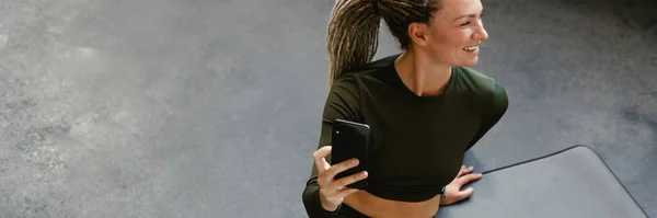 Ung Sportskvinne Smiler Bruker Mobiltelefon Mens Hun Sitter Matta Innendørs – stockfoto