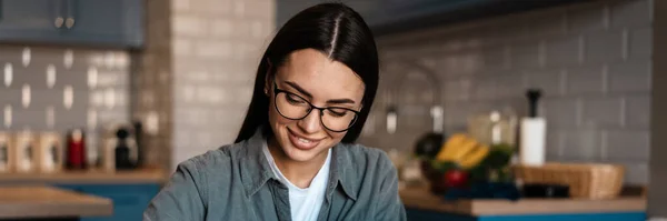 Biała Uśmiechnięta Kobieta Zapisuje Notatki Podczas Pracy Laptopem Domowej Kuchni — Zdjęcie stockowe