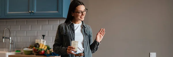 stock image Happy mid aged brunette woman holding glass with beverage while standing at the kitchen table, on a video call via laptop computer, waving hand