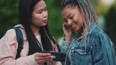 Mutlu çok kültürlü bayan arkadaşlar kulaklıkla müzik dinliyor ve yazları sokakta cep telefonuyla video izliyorlar.