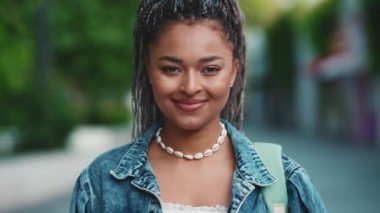 Saçları örgülü, gülümseyen Afrikalı kadın yazın sokakta kameraya bakıyor.