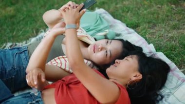 İki güzel Asyalı kadın piknikte çimlerin üzerinde uzanırken cep telefonu kamerasıyla selfie çekiyorlar.