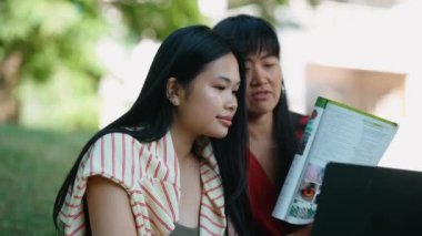 İki sevimli Asyalı kız öğrenci laptopta video ile konuşuyor ve açık havada kitap gösteriyor.