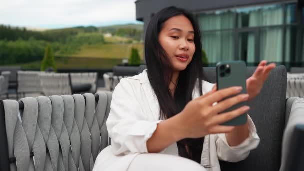 ポジティブなアジアのブルネット女性休憩し 夏のバルコニーの山のリゾートで携帯電話でビデオ通話で話す — ストック動画