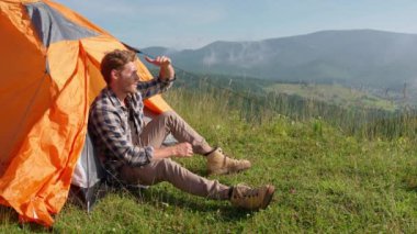Dağlarda çadırda otururken gülümseyen sarışın bir adam manzaraya bakıyor. Kamp kavramı