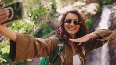 Güneş gözlüklü neşeli turist kadın telefonda dağlarda şelaleye karşı selfie çekiyor.