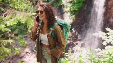 Güneş gözlüklü neşeli turist kadın telefonda dağlarda şelaleye karşı konuşuyor.