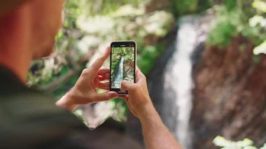 Turistin arkası dağlardaki şelalenin fotoğrafını çekiyor.