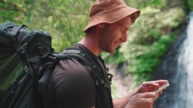 Panama şapkalı, gülümseyen sarışın turist dağlarda şelaleye karşı telefonla mesajlaşıyor.