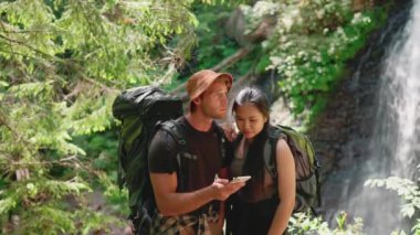 Telefondan güzergaha bakan ve dağlardaki şelaleyi işaret eden mutlu çok uluslu turist çifti.